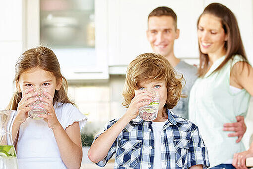Zwei Kinder trinken Wasser aus Gläsern, im Hintergrund ihre Eltern Zwei Kinder trinken Wasser aus Gläsern, im Hintergrund ihre Eltern