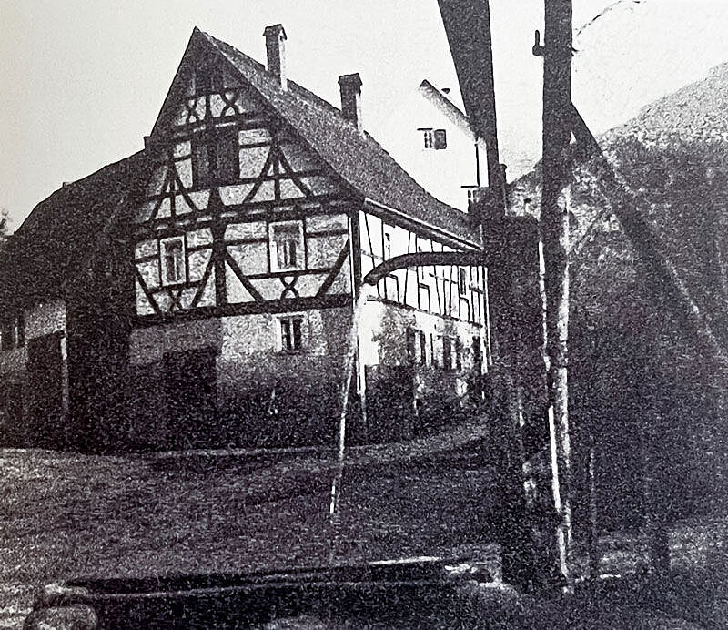 Im Vordergrund ein Brunnen, aus dem Wasser fließt, im Hintergrund ein Fachwerkhaus Im Vordergrund ein Brunnen, aus dem Wasser fließt, im Hintergrund ein Fachwerkhaus