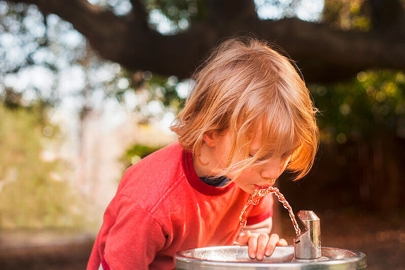 Junge trinkt Wasser an einem Trinkbrunnen. Im Hintergrund ist ein Park zu sehen. Junge trinkt Wasser an einem Trinkbrunnen. Im Hintergrund ist ein Park zu sehen.
