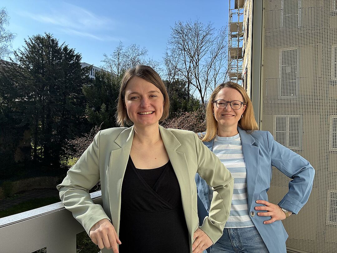 2026_03_Interview_Heck_Volke Zwei Frauen stehen selbstbewusst mit eingestützen Armen auf einem Balkon und schauen freundlich in die Kamera.