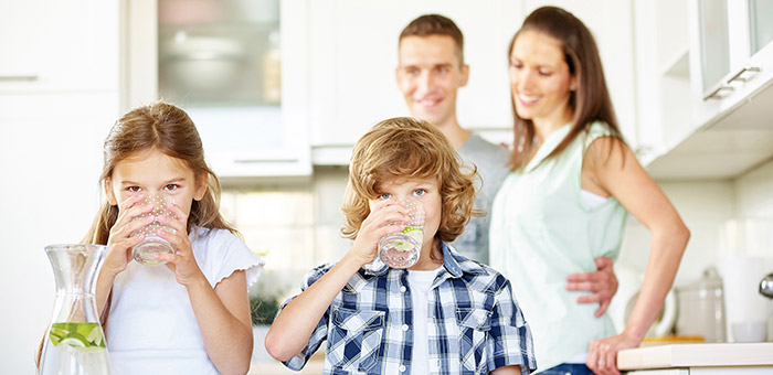 Zwei Kinder trinken Wasser aus Gläsern, im Hintergrund ihre Eltern