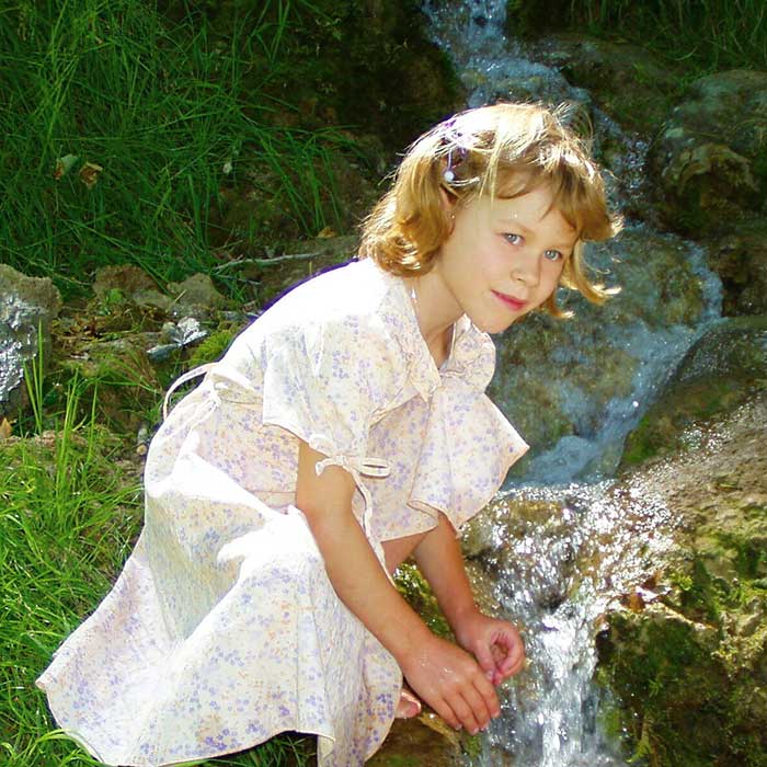Mädchen in einem romantischen Kleid an einem Bachlauf. Es kniet und taucht die Hände in das klare Wasser.